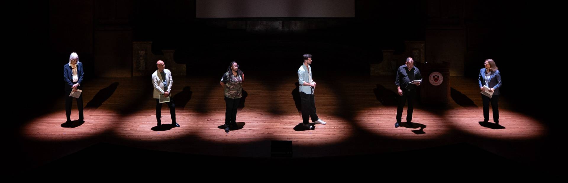 Six people standing under the spotlight onstage.