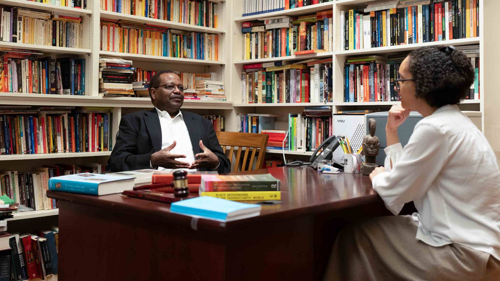 A professor and a student talking in a room full of books.